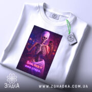 Футболка я вже стара. Біла футболка, складена на поверхні, з принтом скелета у рушнику, що тримає телефон. На футболці є етикетка з зеленим шнурком.