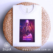 Футболка я вже стара. Біла футболка, складена на плетеній основі, з принтом скелета у рушнику, що тримає телефон. Принт яскравий, фон світлий.