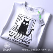 Футболка я з техпідтримки, білого кольору, зображена з верхнього ракурсу, на фоні світло-сірого кольору, принт чорного кольору з котом на ноутбуці та текстом 'НЕ ХВИЛЮЙСЯ Я З ТЕХПІДТРИМКИ', з етикеткою зеленого кольору.