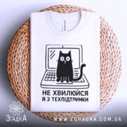 Футболка я з техпідтримки, білого кольору, зображена з верхнього ракурсу на плетеному кошику, фон світло-сірий, принт чорного кольору з котом на ноутбуці та текстом 'НЕ ХВИЛЮЙСЯ Я З ТЕХПІДТРИМКИ'.