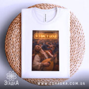 Футболка я з вас у шоці. Біла футболка, розкладена на плетеній основі, з принтом чоловіка, що грає на бандурі. На фоні видно людей у залі, текст 'Я З ВАС У ШОЩ!' яскраво підсвічений.