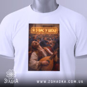Футболка я з вас у шоці. Біла футболка, складена на поверхні, з принтом чоловіка, що грає на бандурі. На фоні видно людей у залі, текст 'Я З ВАС У ШОЩ!' виділяється яскраво.