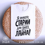 Футболка я занадто старий. Біла футболка, складена на плетеній кошику, з видимим ярликом. Фон світлий, з легким градієнтом. Принт чорного кольору, розташований по центру, з великими літерами.