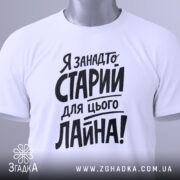 Футболка я занадто старий. Біла футболка, складена на поверхні, з видимим ярликом. Фон світлий, з легким градієнтом. Принт чорного кольору, розташований по центру, з великими літерами.