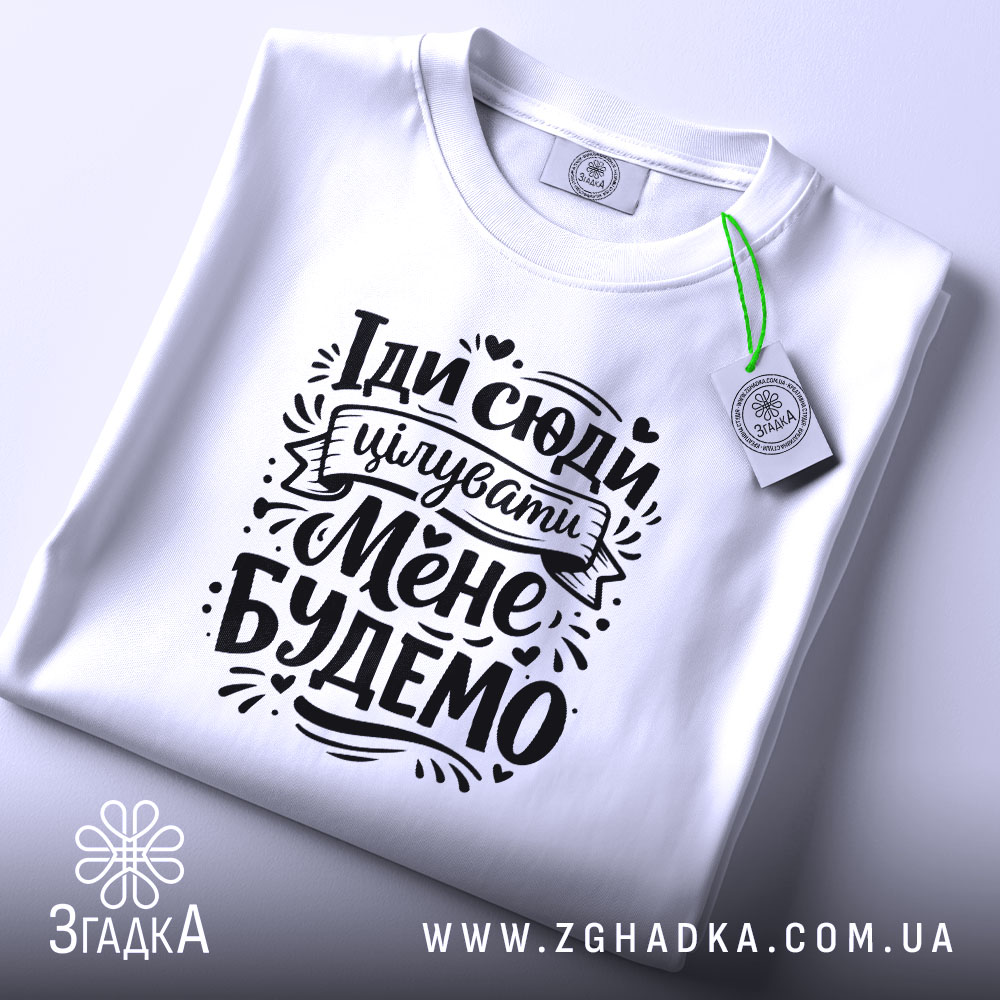 Футболка Іди сюди цілувати біла, складена на столі, фон світло-сірий, принт чорного кольору з текстом "Іди сюди цілувати Мене Будемо" з елементами декору, на комірці зелена етикетка.