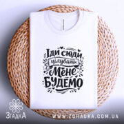 Футболка Іди сюди цілувати біла, складена на плетеній основі, фон світло-сірий, принт чорного кольору з текстом "Іди сюди цілувати Мене Будемо" оформлений у стилі рукописного шрифту.