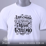 Футболка Іди сюди цілувати біла, складена на столі, фон світло-сірий, принт чорного кольору з текстом "Іди сюди цілувати Мене Будемо" з елементами декору, на комірці зелена етикетка.