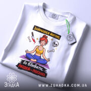 Футболка йога чорний гумор, біла, складена на столі, фон світло-сірий, принт з дівчиною в позі йоги, червоним волоссям, жовтою майкою та синіми штанами, прикріплена етикетка з логотипом.