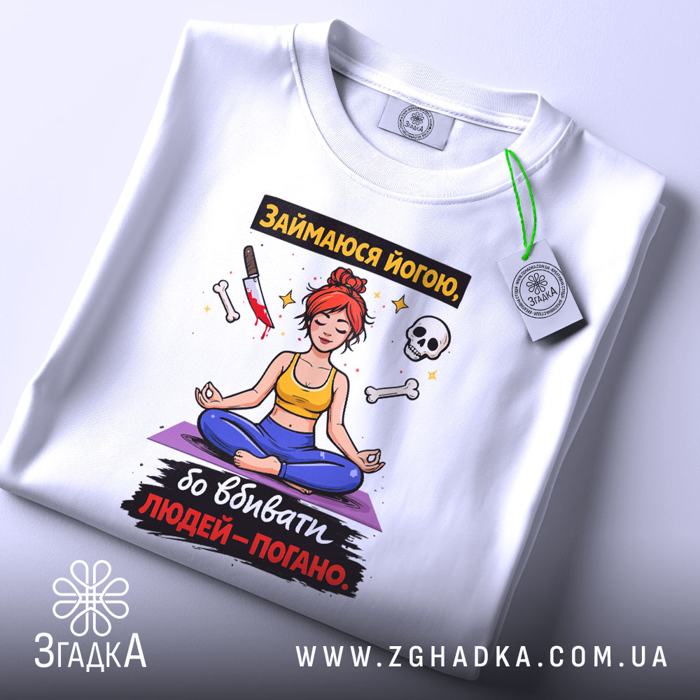 Футболка йога чорний гумор, біла, складена на столі, фон світло-сірий, принт з дівчиною в позі йоги, червоним волоссям, жовтою майкою та синіми штанами, прикріплена етикетка з логотипом.
