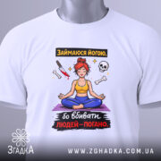 Футболка йога чорний гумор, біла, складена на столі, фон світло-сірий, принт з дівчиною в позі йоги, червоним волоссям, жовтою майкою та синіми штанами, прикріплена етикетка з логотипом.