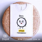 Футболка його краща половинка, білого кольору, складена на плетеній основі, фон світлий, принт з малюнком круглого персонажа з червоними губами та написом 'КРАЩА ПОЛОВИНКА' жовтого кольору, видно деталі тканини.