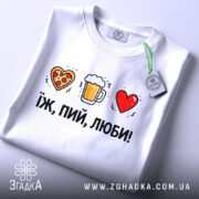 Футболка їж пий люби білого кольору, складена на поверхні, фон світло-сірий, принт з малюнками серця з піцою, пива в кружці та червоного серця, текст чорний, розташований під малюнками, на комірці є етикетка.