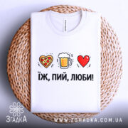 Футболка їж пий люби білого кольору, розкладена на плетеній основі, фон світло-сірий, принт з малюнками серця з піцою, пива в кружці та червоного серця, текст чорний, розташований під малюнками.