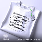 Футболка хороші люди, біла, складена на столі, фон світло-сірий, принт чорного кольору з текстом та зірочками, на комірці етикетка.