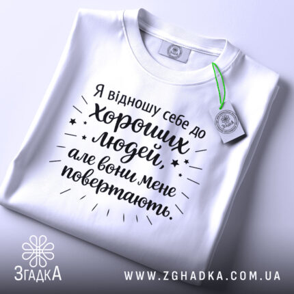 Футболка хороші люди, біла, складена на столі, фон світло-сірий, принт чорного кольору з текстом та зірочками, на комірці етикетка.