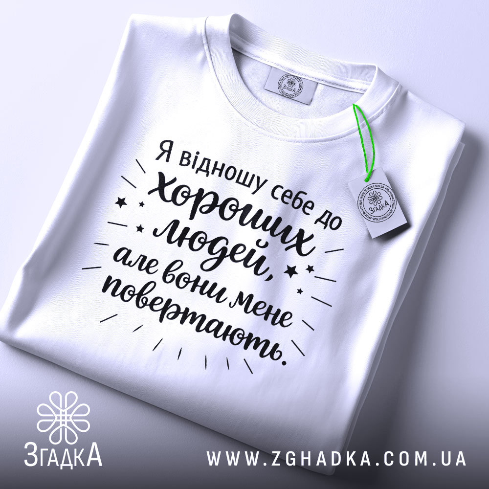 Футболка хороші люди, біла, складена на столі, фон світло-сірий, принт чорного кольору з текстом та зірочками, на комірці етикетка.