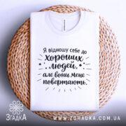 Футболка хороші люди, біла, складена на плетеній кошику, фон світло-сірий, принт чорного кольору з текстом та зірочками.