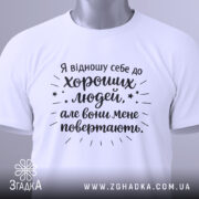 Футболка хороші люди, біла, складена на столі, фон світло-сірий, принт чорного кольору з текстом та зірочками, на комірці етикетка.