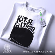 Футболка Кіт я тиша. Біла футболка з коротким рукавом, розкладена на поверхні. Принт чорного кольору з малюнком кота та текстом на українській мові видно чітко. На футболці є етикетка з зеленим шнурком. Фон світлий, однорідний.