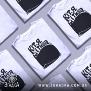 Футболка Кіт я тиша. Біла футболка з коротким рукавом, зображена на манекені з блискучою поверхнею. Принт чорного кольору з малюнком кота та текстом на українській мові розташований по центру. Фон нейтральний, світлий.