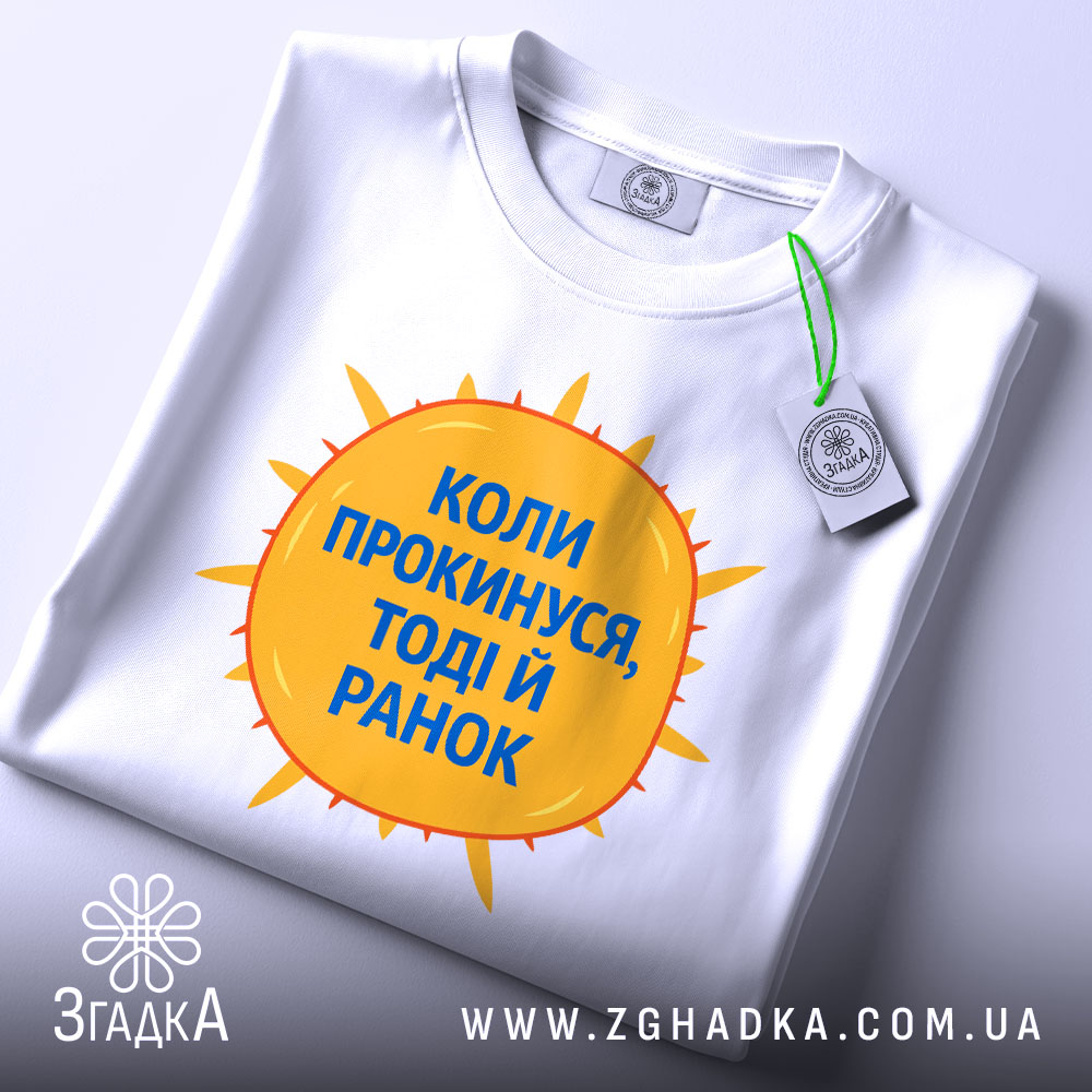 Футболка коли прокинувся: складена біла футболка з принтом у вигляді сонця жовтого кольору і текстом синього кольору "Коли прокинусь, тоді й ранок", фон світло-сірий, ракурс зверху.