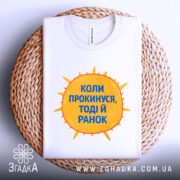 Футболка коли прокинувся: складена біла футболка на плетеній основі, з принтом у вигляді сонця жовтого кольору і текстом синього кольору "Коли прокинусь, тоді й ранок", фон світло-сірий, ракурс зверху.