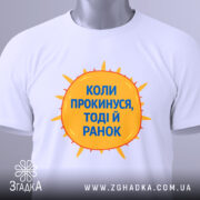 Футболка коли прокинувся: складена біла футболка з принтом у вигляді сонця жовтого кольору і текстом синього кольору "Коли прокинусь, тоді й ранок", фон світло-сірий, ракурс зверху.