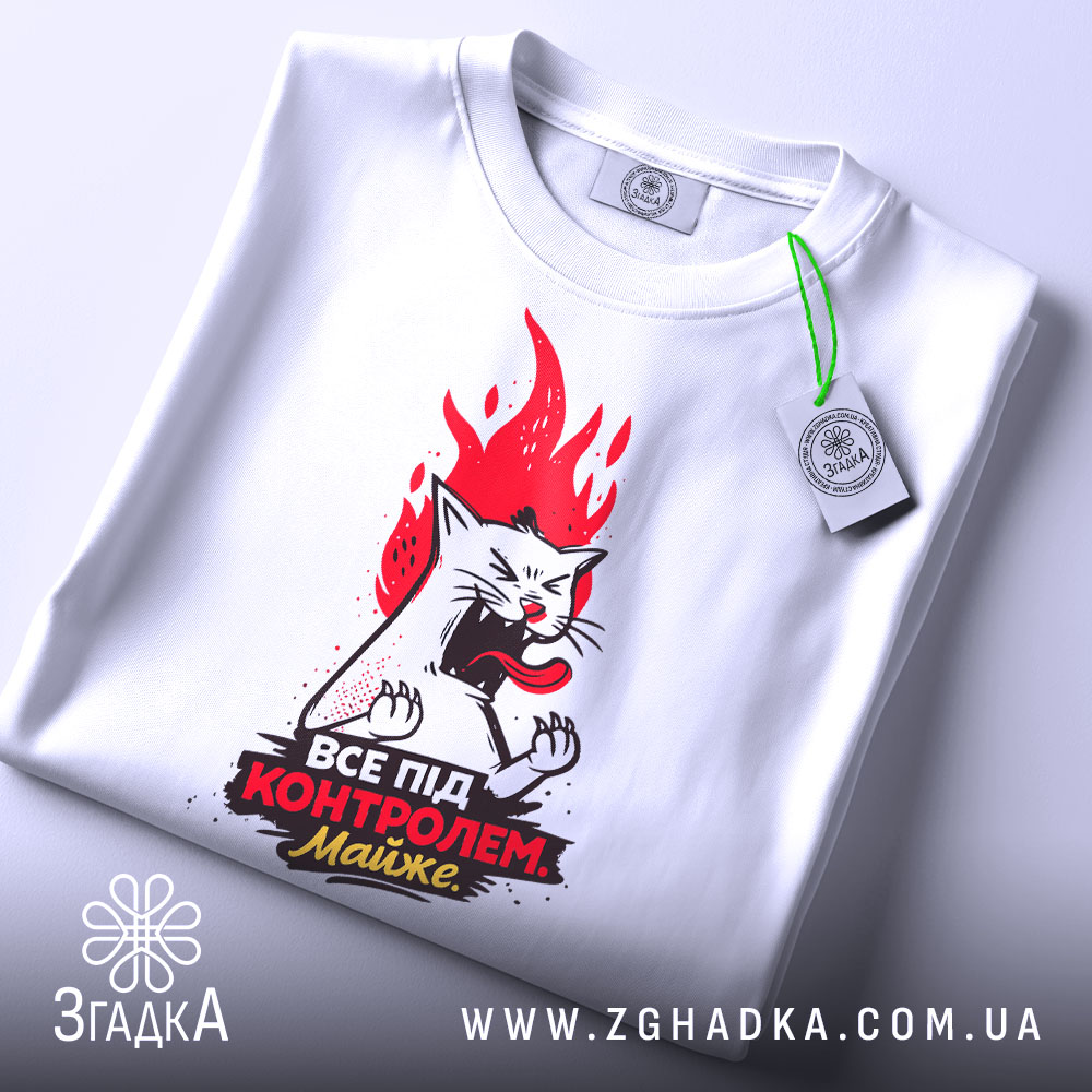 Футболка контроль без паніки. Біла футболка, складена на столі, з видимим принтом кота з червоним полум'ям. На футболці є етикетка з зеленим шнурком.
