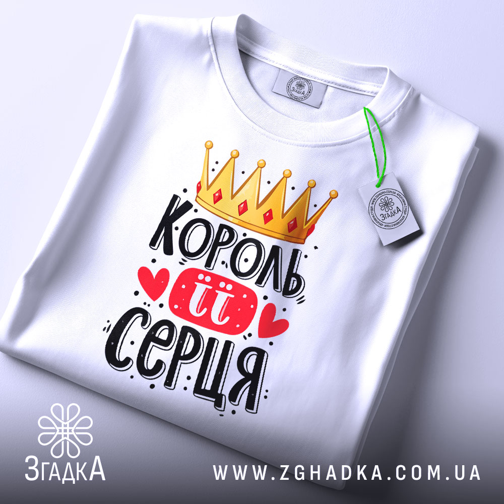 Футболка король її серця, білого кольору, складена на столі, фон світло-сірий, принт з короною, червоними сердечками та написом чорного кольору, з зеленим ярликом.