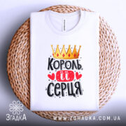 Футболка король її серця, білого кольору, складена на плетеному кошику, фон світло-сірий, принт з короною, червоними сердечками та написом чорного кольору.