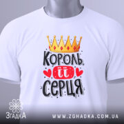 Футболка король її серця, білого кольору, складена на столі, фон світло-сірий, принт з короною, червоними сердечками та написом чорного кольору, з зеленим ярликом.