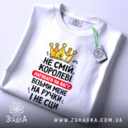 Футболка королева без страху, біла, складена на поверхні, фон світлий, принт з жовтою короною та текстом червоного і чорного кольорів, з етикеткою зеленого кольору.