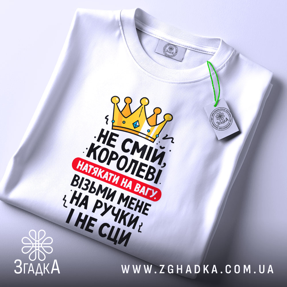 Футболка королева без страху, біла, складена на поверхні, фон світлий, принт з жовтою короною та текстом червоного і чорного кольорів, з етикеткою зеленого кольору.