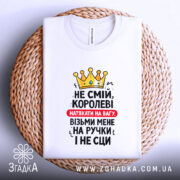 Футболка королева без страху, біла, складена на плетеній основі, фон світлий, принт з жовтою короною та текстом червоного і чорного кольорів.