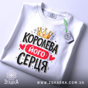 Футболка королева його серця, біла, складена на столі, фон світло-сірий, принт з короною, червоним написом і сердечками, з етикеткою на зеленому шнурку.