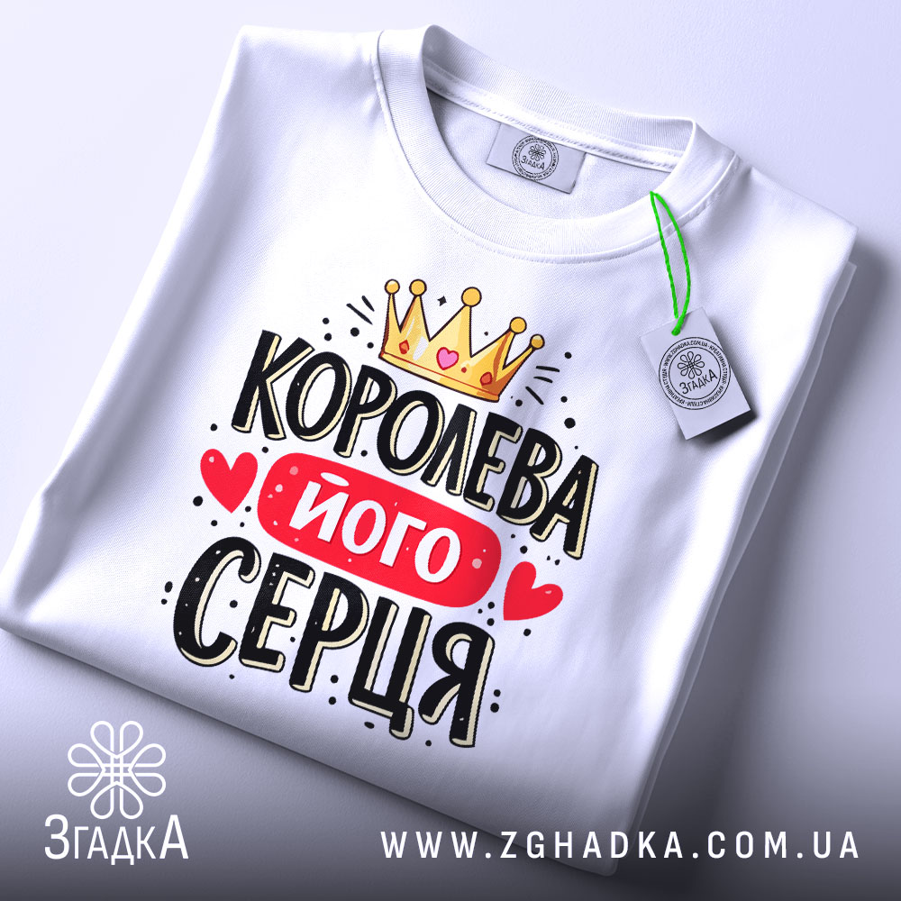 Футболка королева його серця, біла, складена на столі, фон світло-сірий, принт з короною, червоним написом і сердечками, з етикеткою на зеленому шнурку.