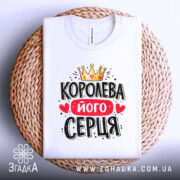 Футболка королева його серця, біла, складена на плетеній кошику, фон світло-сірий, принт з короною, червоним написом і сердечками.