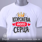 Футболка королева його серця, біла, складена на столі, фон світло-сірий, принт з короною, червоним написом і сердечками, з етикеткою на зеленому шнурку.