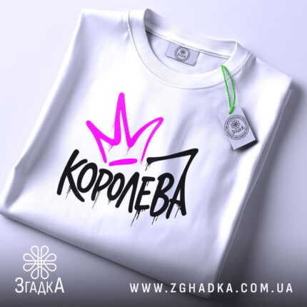 Футболка королева білого кольору, складена на столі, з видимим етикеткою, фон світло-сірий, принт з фіолетовою короною та чорним написом 'КОРОЛЕВА' з крапельками.