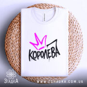 Футболка королева білого кольору, складена на плетеній кошику, фон світло-сірий, принт з фіолетовою короною та чорним написом 'КОРОЛЕВА' з крапельками.