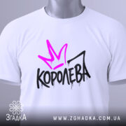 Футболка королева білого кольору, складена на столі, з видимим етикеткою, фон світло-сірий, принт з фіолетовою короною та чорним написом 'КОРОЛЕВА' з крапельками.