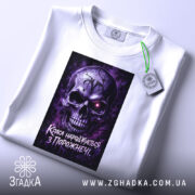 Футболка краса з порожнечі, біла, зображена в горизонтальному положенні, фон світлий, принт з черепом у фіолетових тонах, на футболці є етикетка з зеленим шнурком.
