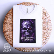 Футболка краса з порожнечі, біла, зображена в горизонтальному положенні на плетеній основі, фон світлий, принт з черепом у фіолетових тонах, напис на футболці чітко видимий.