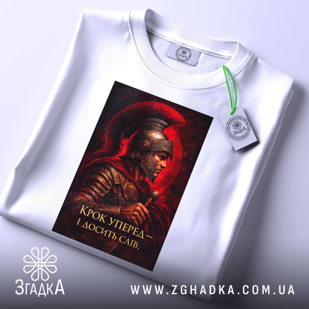 Футболка крок уперед. Біла футболка, зображена зверху, принт з воїном в шоломі на червоному фоні, текст: 'Крок уперед – і досягти слави', з зеленим ярликом на комірці.