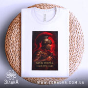 Футболка крок уперед. Біла футболка, складена на плетеній кошику, принт з воїном в шоломі на червоному фоні, текст: 'Крок уперед – і досягти слави', вид з верхньої частини.