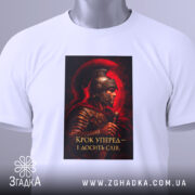 Футболка крок уперед. Біла футболка, зображена зверху, принт з воїном в шоломі на червоному фоні, текст: 'Крок уперед – і досягти слави', з зеленим ярликом на комірці.
