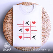 Футболка любов завжди перемагає, складена футболка білого кольору на плетеній основі, принт у стилі гри "Хрестики-нулики" з червоними серцями та чорними "Х", фон світло-блакитний, ракурс зверху.