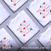 Футболка любов завжди перемагає, біла футболка з принтом у стилі гри "Хрестики-нулики", на грудях зображені червоні серця та чорні "Х", фон світло-сірий, ракурс фронтальний.