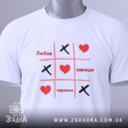 Футболка любов завжди перемагає, складена футболка білого кольору з принтом у стилі гри "Хрестики-нулики", на ній видно червоні серця та чорні "Х", фон світло-блакитний, ракурс зверху.