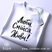 Футболка люби смійся живи, білого кольору, складена на плоскій поверхні, фон світло-сірий, принт чорного кольору з написом 'Люби. Смійся. Живи!' у стилі рукописного шрифту, з етикеткою зеленого кольору.